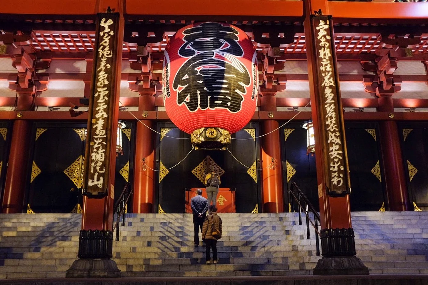 Two visitors stand before the illuminated main hall of Sensoji Temple at night, the great red lantern glowing above them